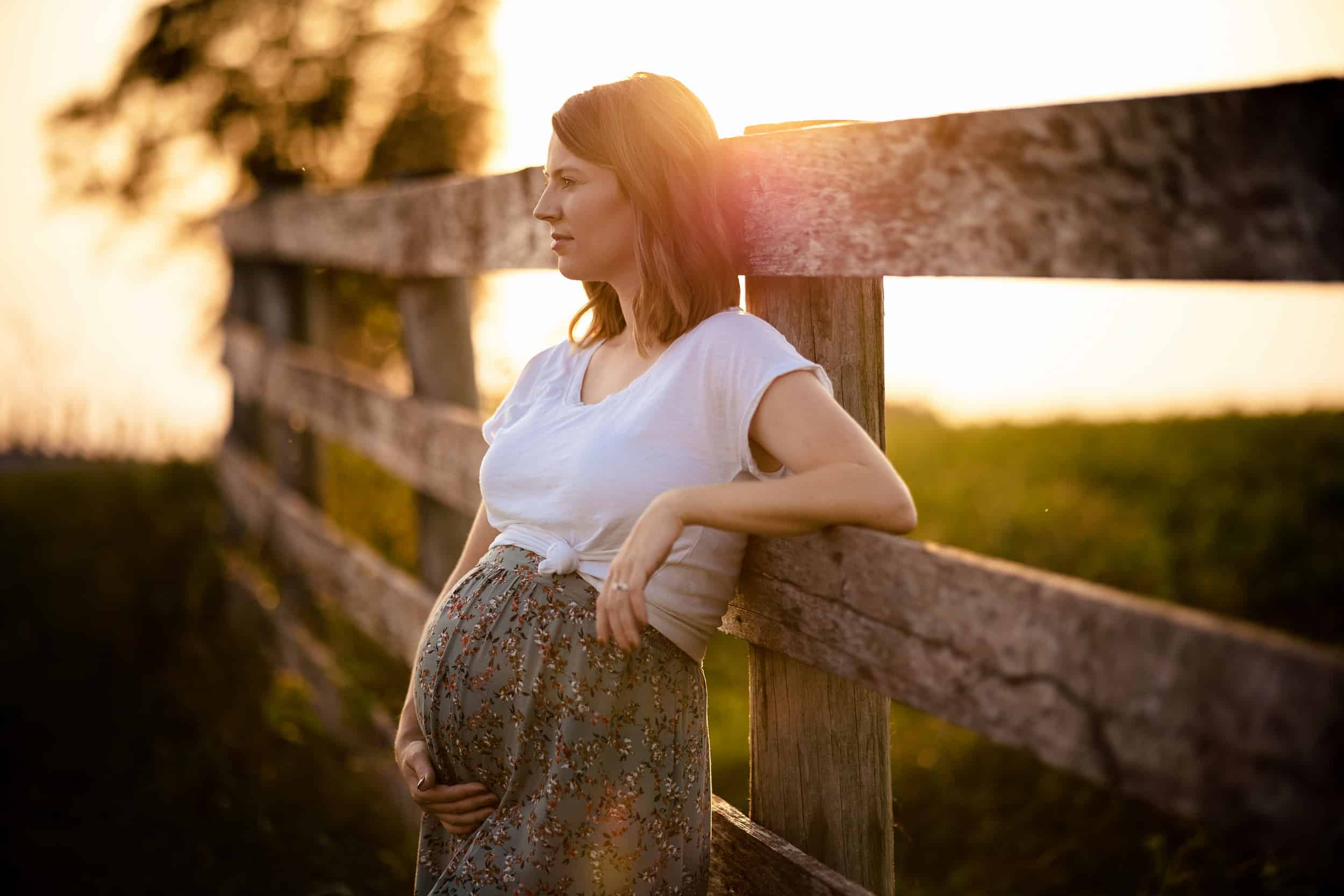A pregnant woman leaning against a fence at sunset.