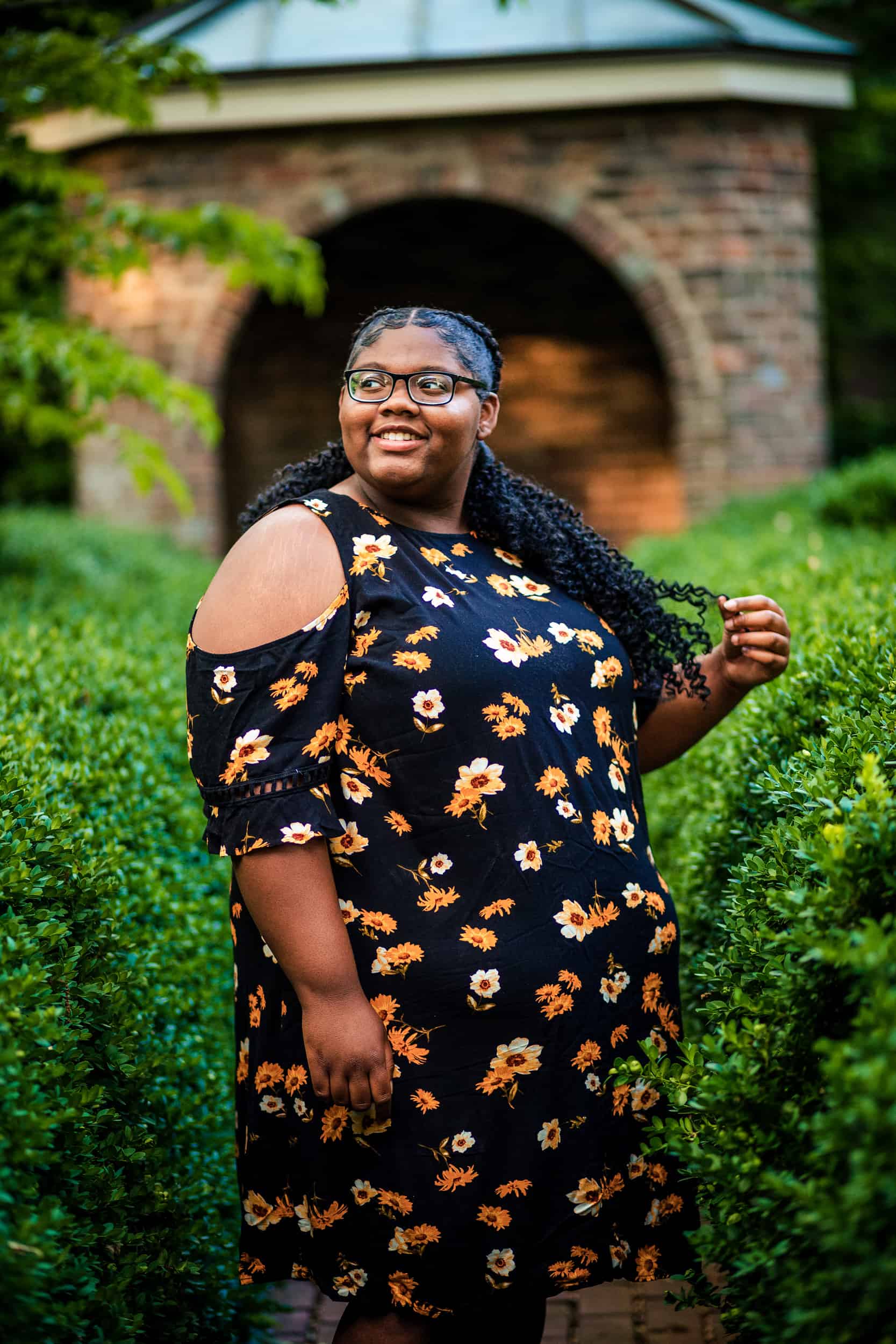 A black woman in a floral dress smiles in front of bushes.