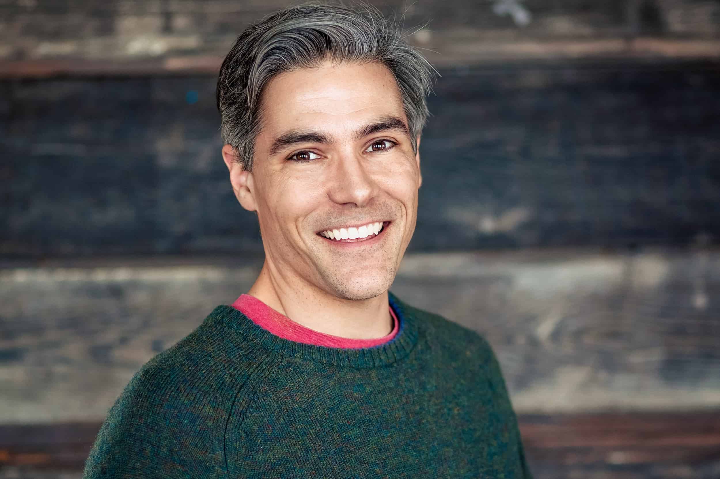 A man in a green sweater smiles in front of a wooden wall.