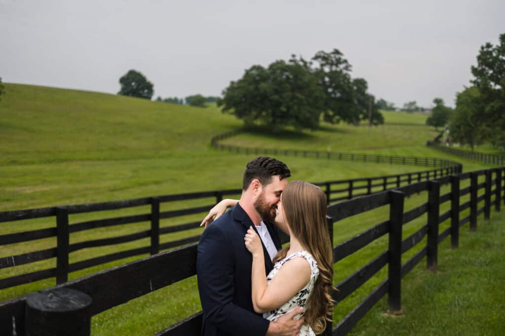 A couple kisses during their Frankfort KY engagement photos in front of a fence in a field.