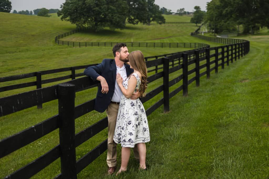 A couple kisses during their Frankfort KY engagement photos in front of a fence in a field.