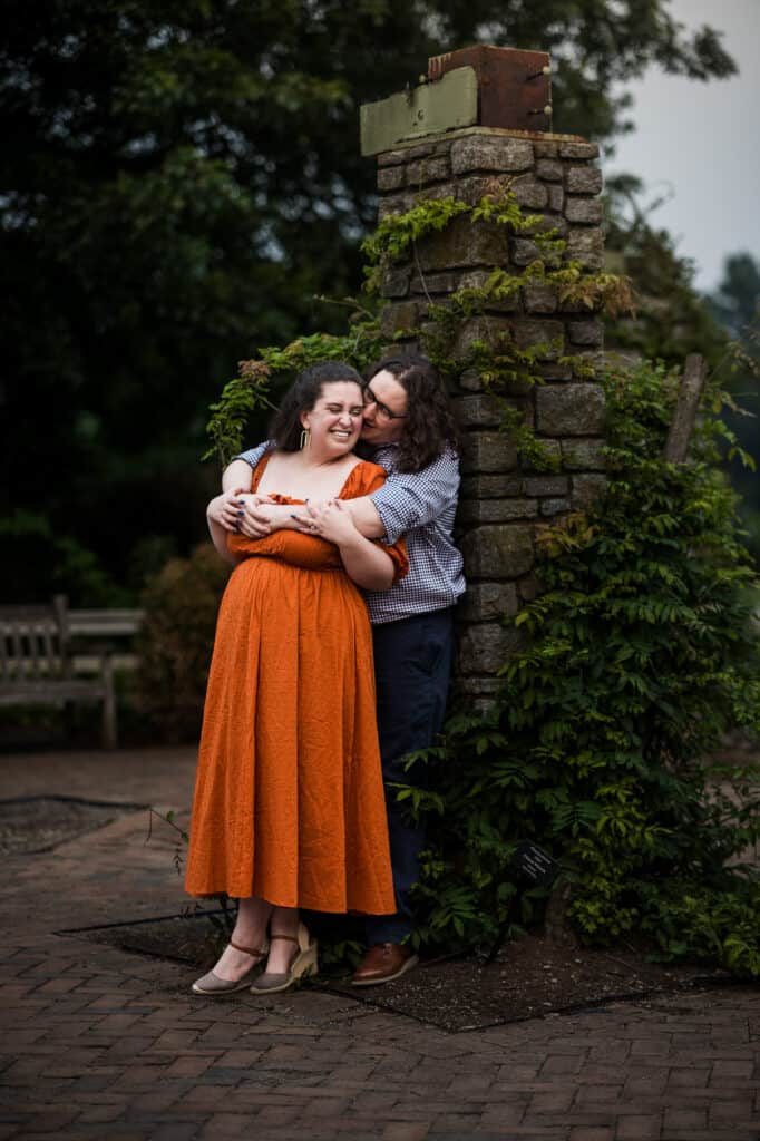 A couple embracing during their engagement session at the UK Arboretum, with a stone wall as the backdrop.