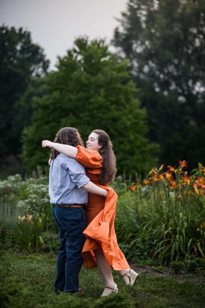 A couple in their engagement session wearing orange dresses, hugging in the beautiful UK Arboretum garden.