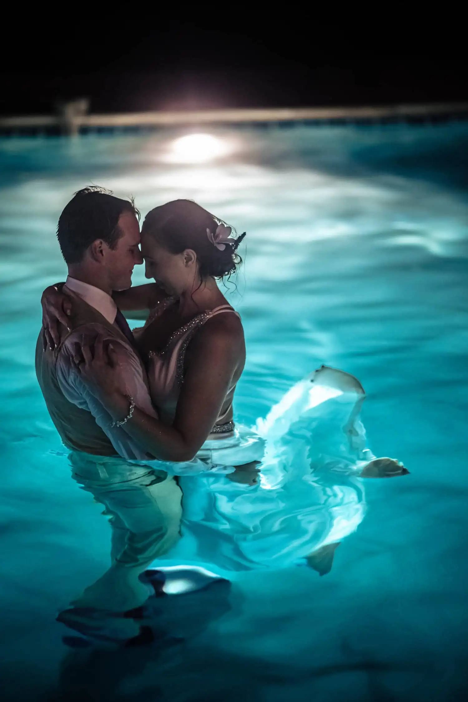 A bride and groom embracing in a pool at night.