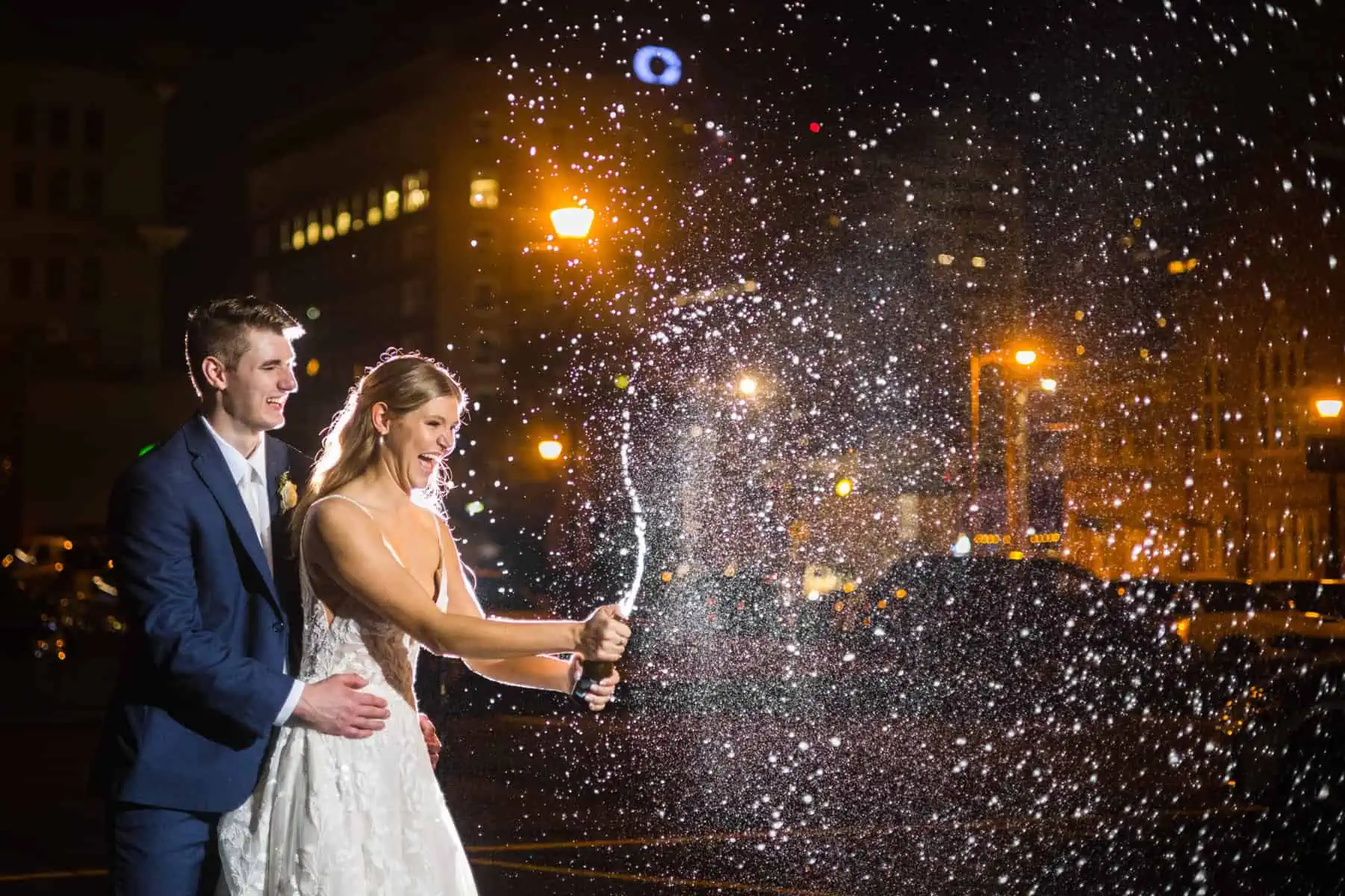 A bride and groom are holding a champagne bottle in front of a building at night.