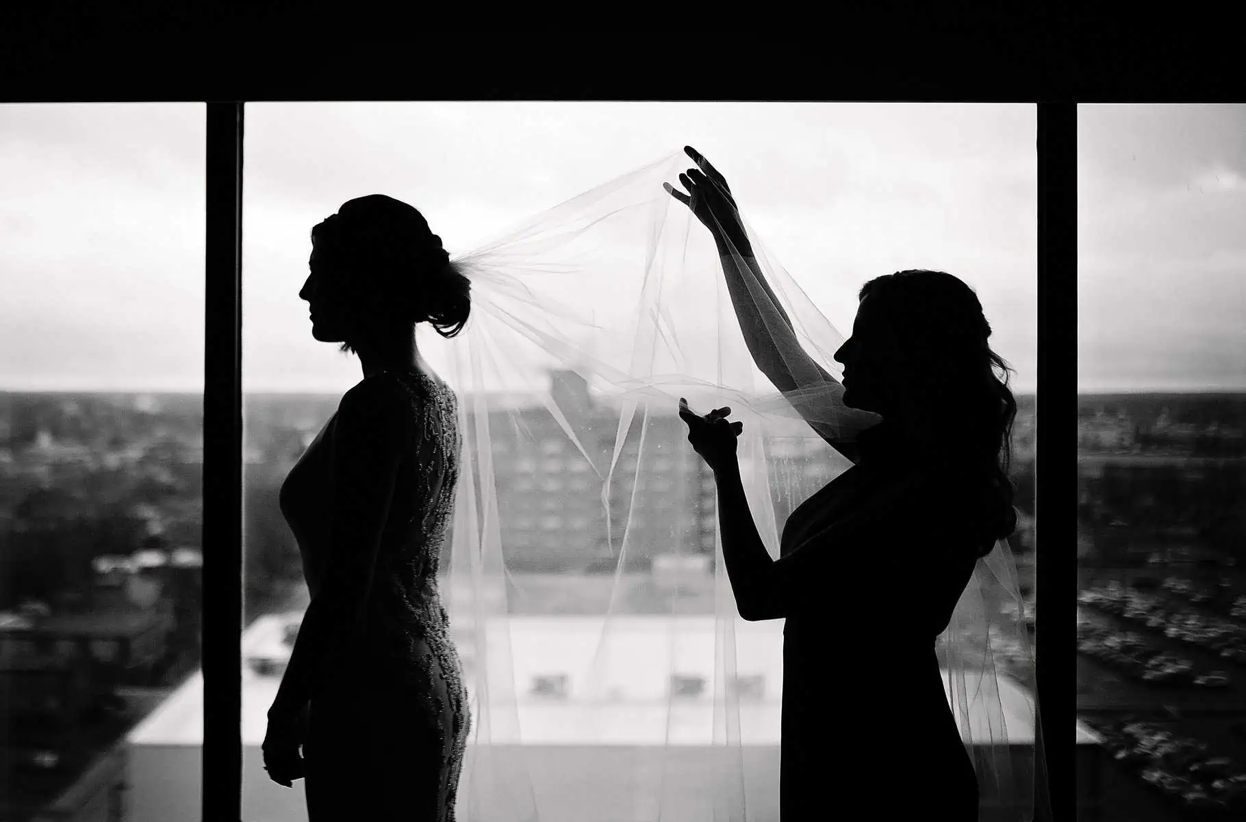 A bride is putting her veil on in front of a window.