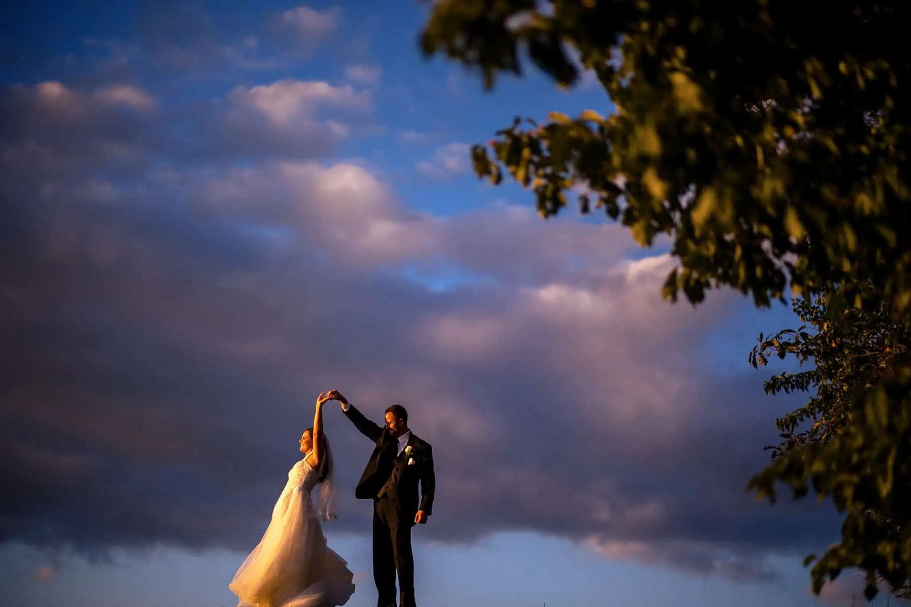 A bride and groom standing on top of a hill under a cloudy sky.