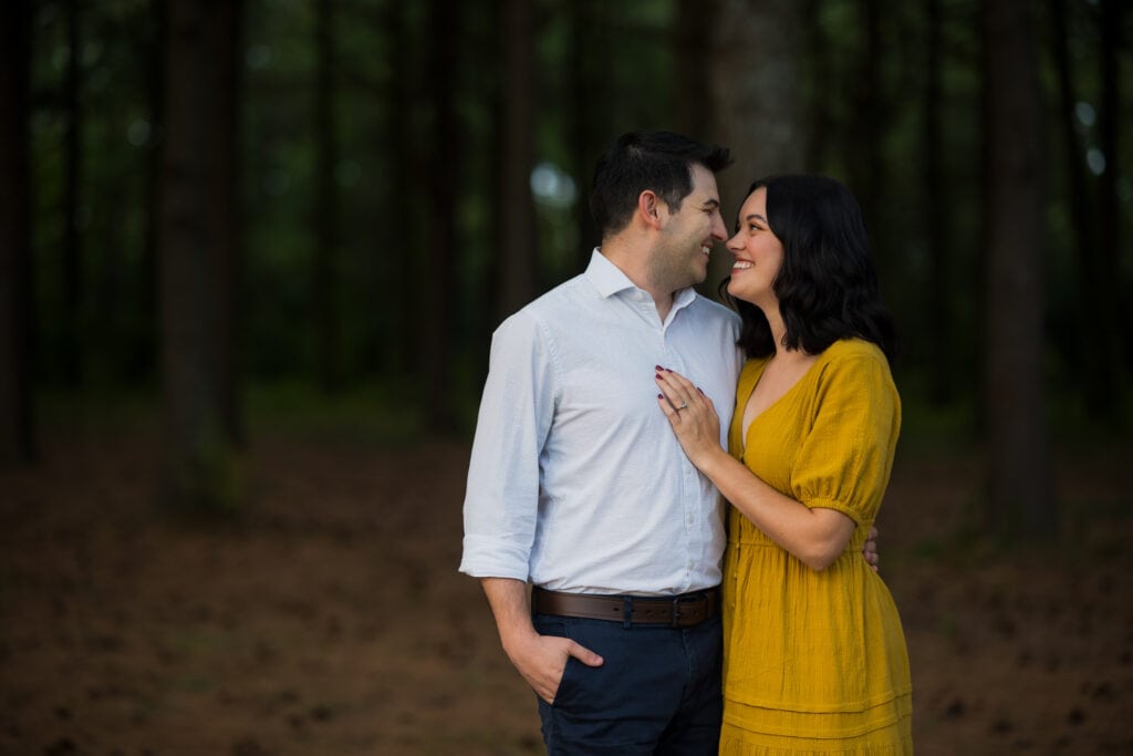 Jacobson Park Engagement Photos Of A Couple In A Yellow Dress Standing In A Wooded Area.