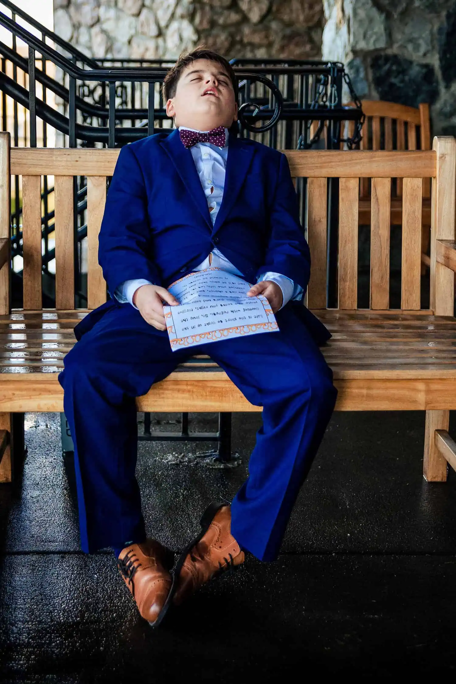 A boy in a blue suit sitting on a wooden bench.