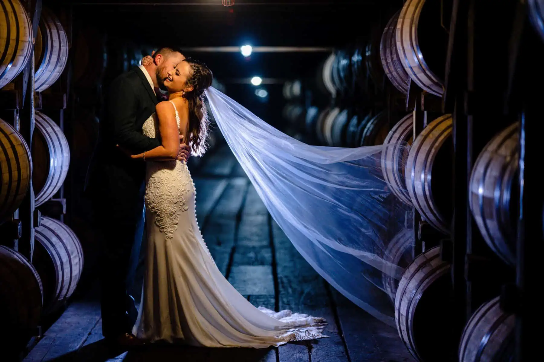A bride and groom kiss in front of barrels of wine.