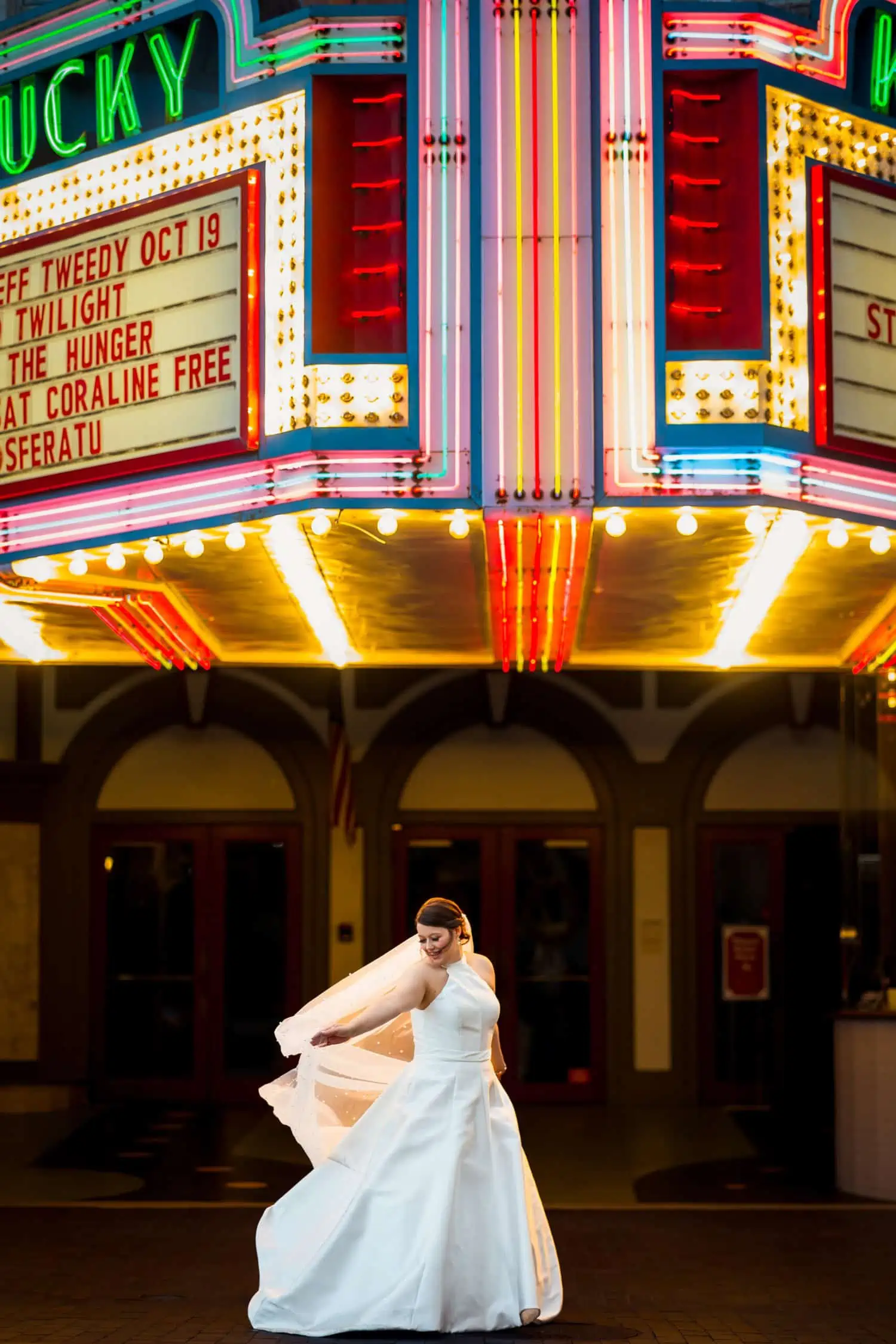 A bride is standing in front of a neon theater sign.