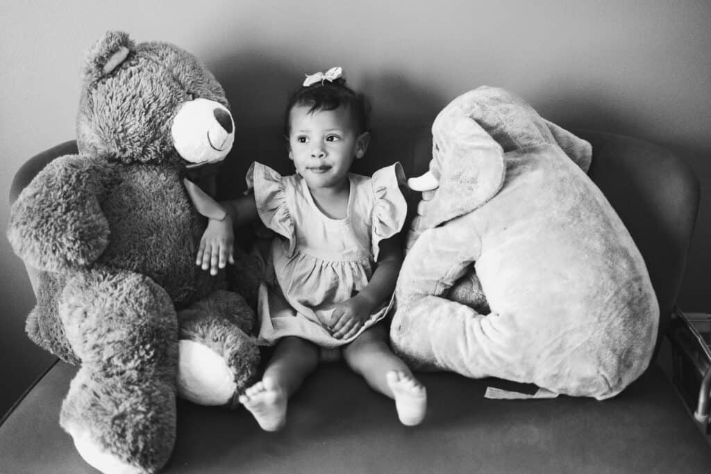 A newborn twin baby girl sits on a couch with two teddy bears, captured beautifully in Lexington's twin photography session.