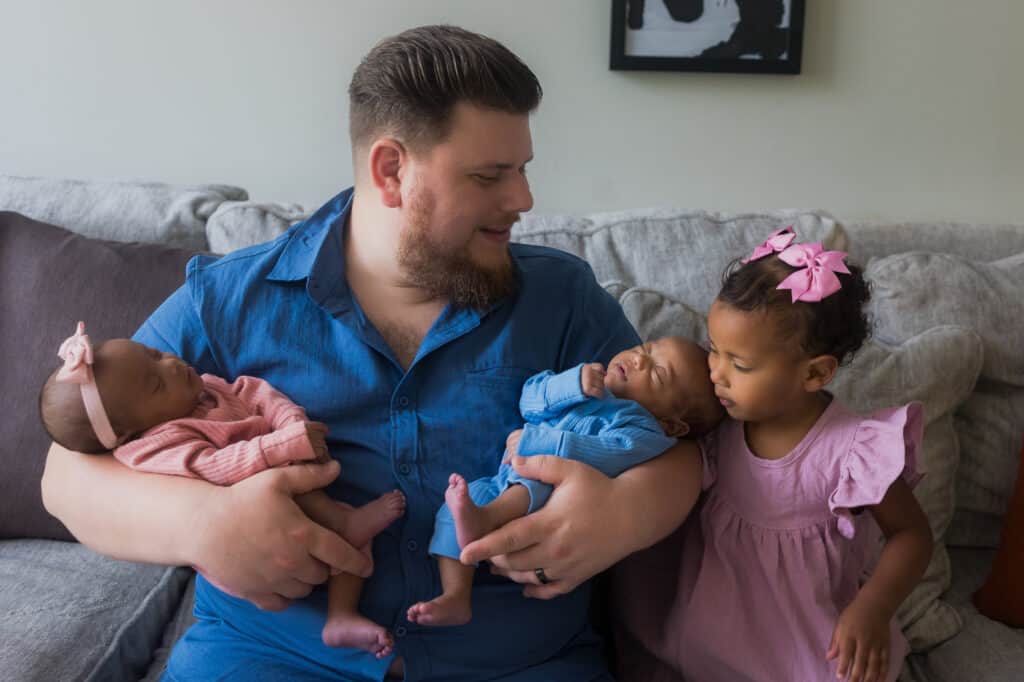 A man delicately cradles two newborn babies on a comfortable couch, creating an enchanting moment captured by professional twin photography in Lexington.