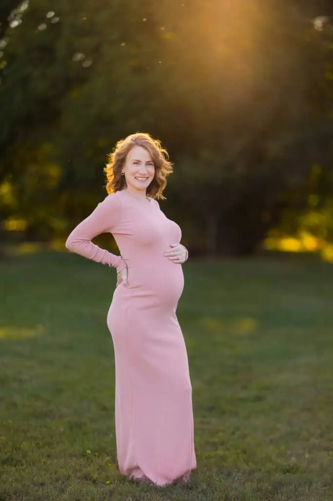 A pregnant woman in a pink dress standing in a field at sunset, captured by a Lexington KY maternity photographer.