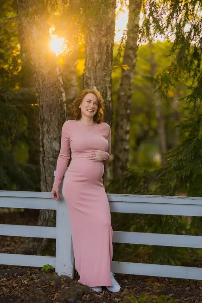 A pregnant woman in a pink dress leaning against a fence captured by Lexington KY Maternity Photographer.