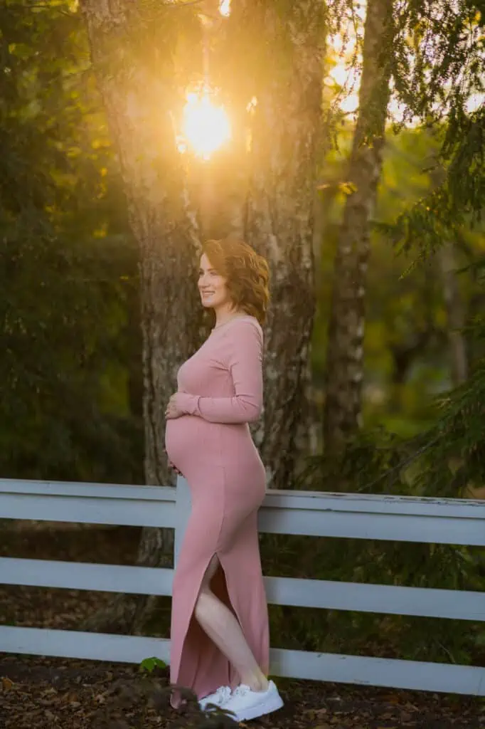 A pregnant woman in a pink dress leaning against a fence in Lexington KY.