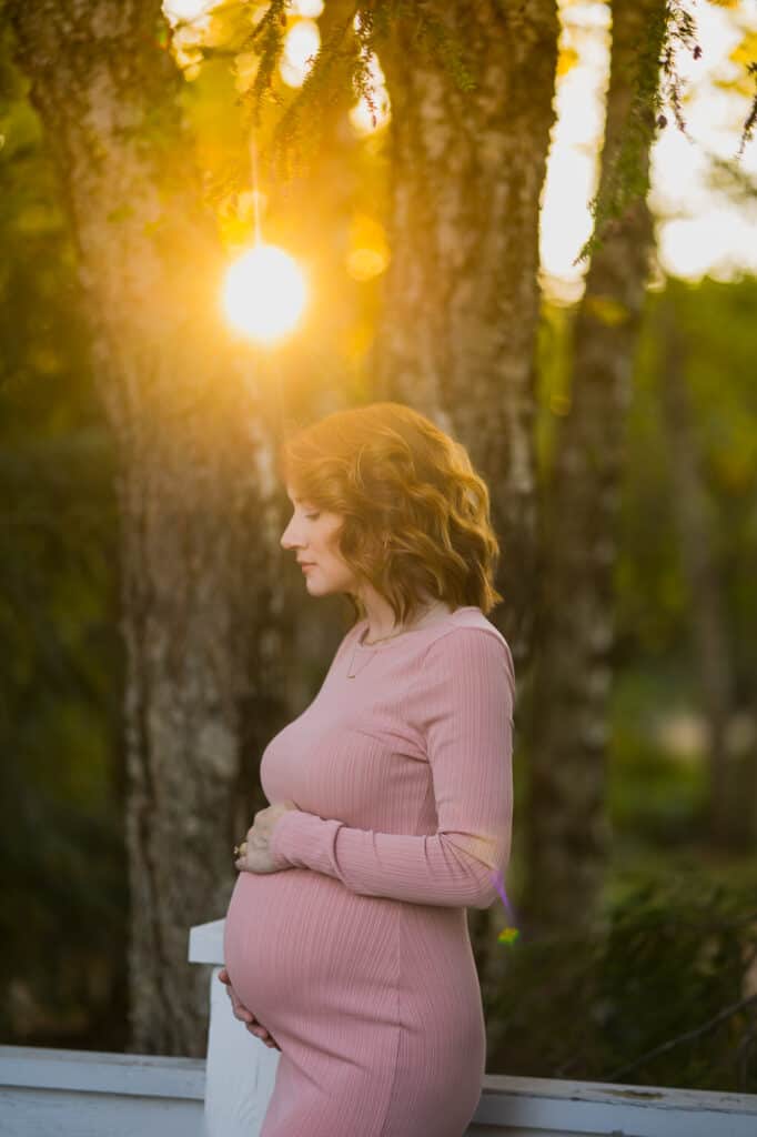 A pregnant woman in a pink dress standing in front of trees at sunset, captured by a Lexington KY maternity photographer.
