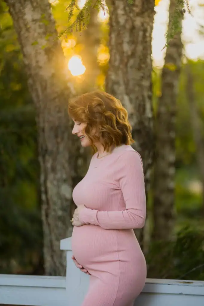 A pregnant woman in a pink dress standing in front of trees captured by a Lexington KY Maternity Photographer.