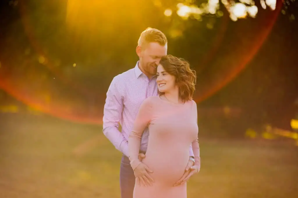 A pregnant couple in Lexington, KY standing in a field with the sun shining down on them as captured by a maternity photographer.