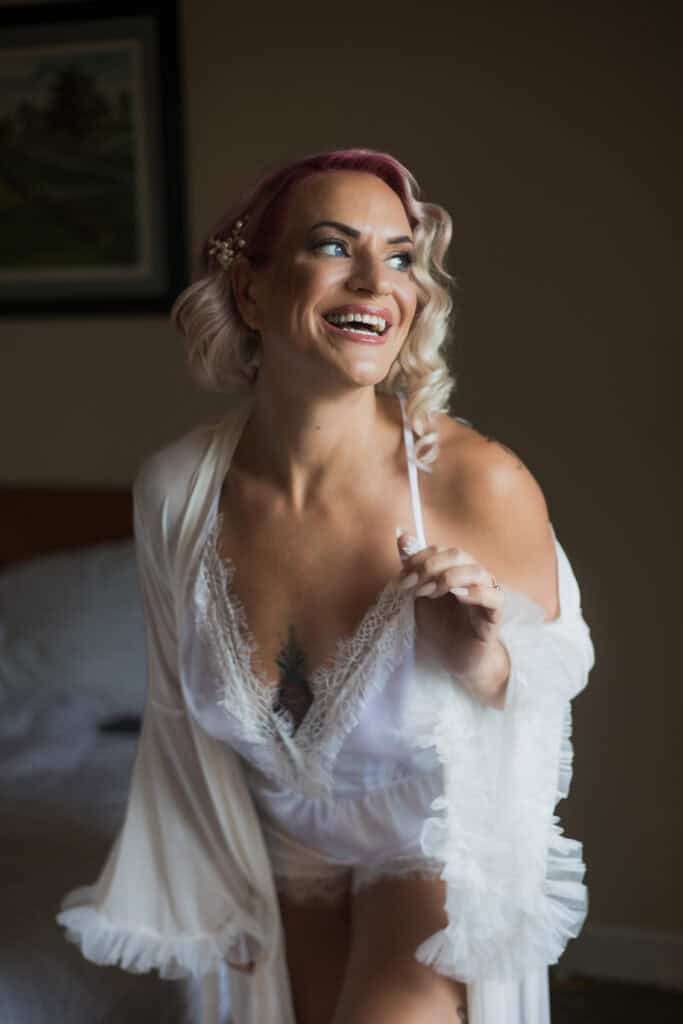 A woman with pink hair, smiling joyfully in a white lace wedding robe, stands in a softly lit hotel room. Capital Plaza in Frankfort, Ky