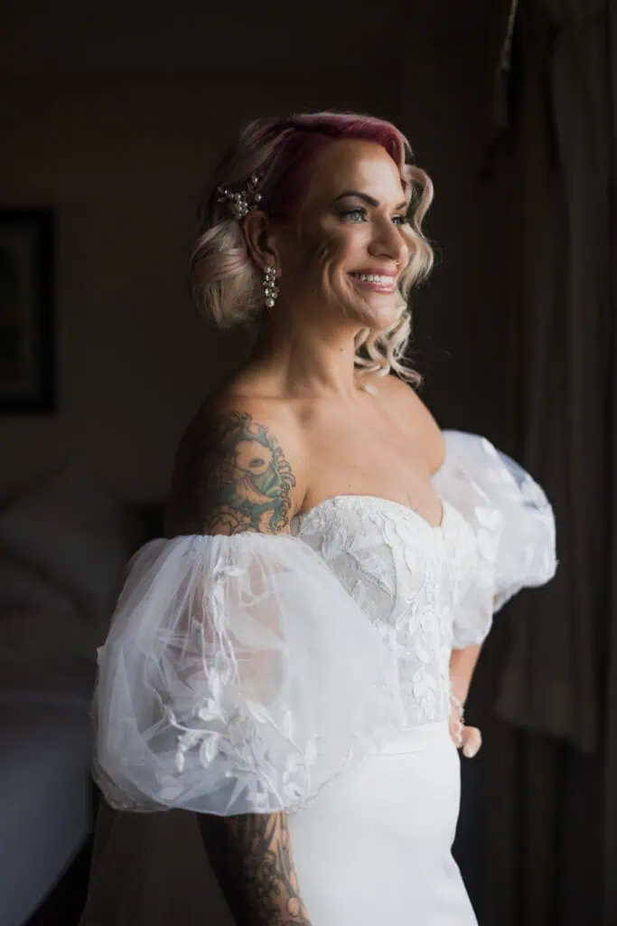 A woman with pink hair and tattoos, wearing a white wedding dress with puffed sleeves, smiles while looking to her side.