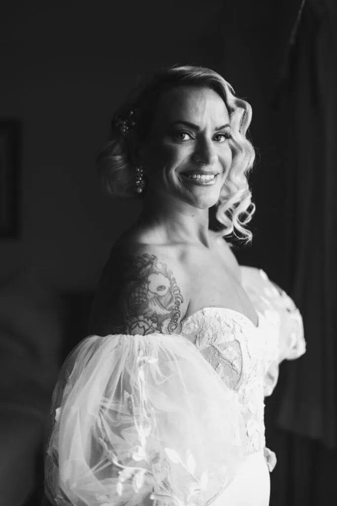 A joyful bride in a white dress with shoulder puff sleeves, showcasing a large tattoo on her left arm, posing in a black and white portrait. Capital Plaza in Frankfort, Ky