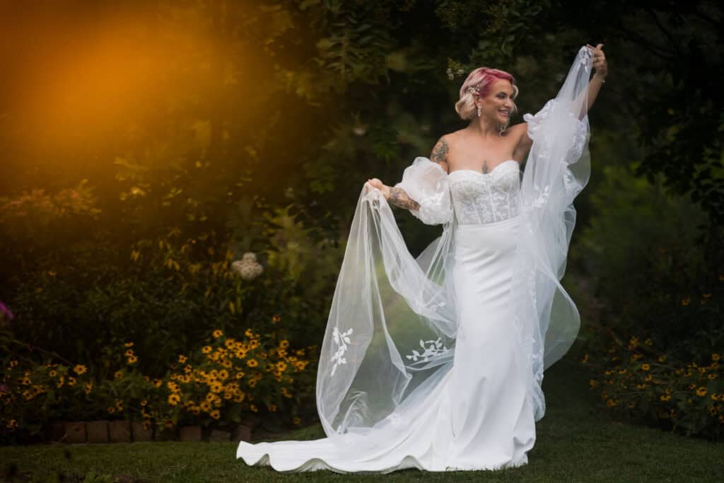 A bride with pink hair smiles while twirling her long veil in a sunlit garden full of sunflowers. Capital Plaza in Frankfort, Ky