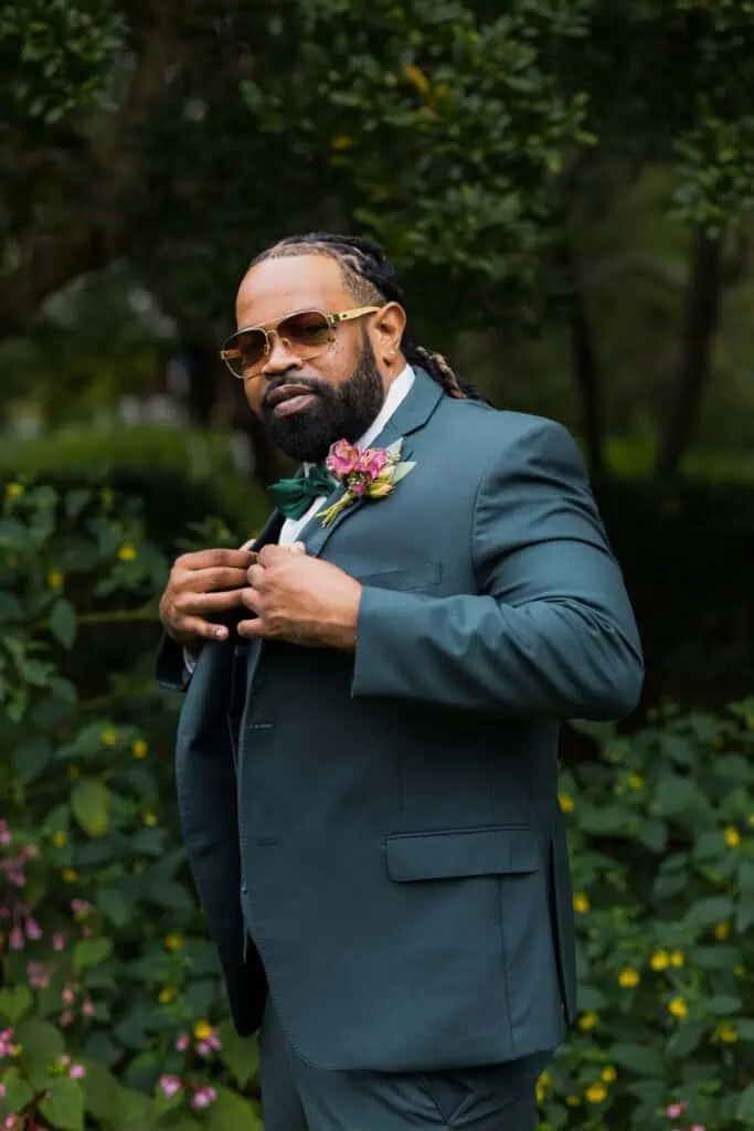 A man in a teal suit adjusts his tie in lush garden, wearing stylish glasses and sporting dreadlocks with a floral boutonniere. Capital Plaza in Frankfort, Ky