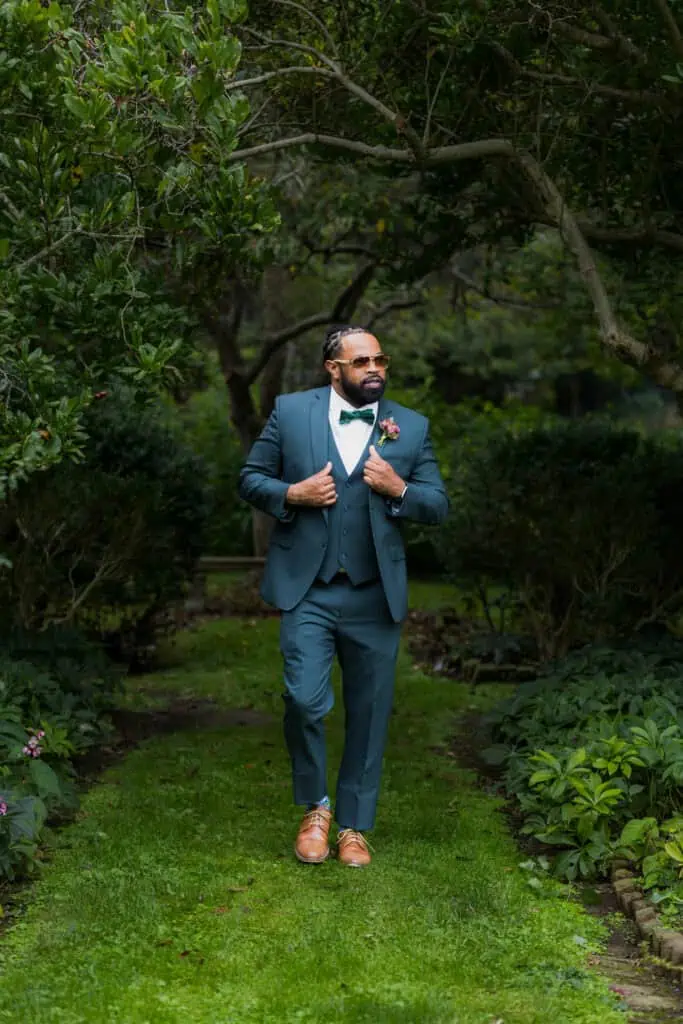 A man in a tailored blue suit and brown shoes poses confidently in a lush garden, adjusting his lapels and wearing sunglasses. Capital Plaza in Frankfort, Ky