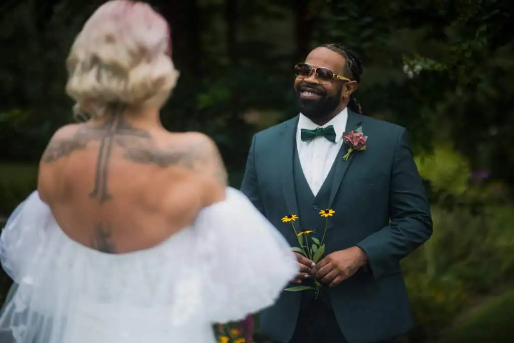 A man in a green suit and bow tie smiling at a woman in a white wedding dress, holding flowers, in a lush garden setting. Capital Plaza in Frankfort, Ky
