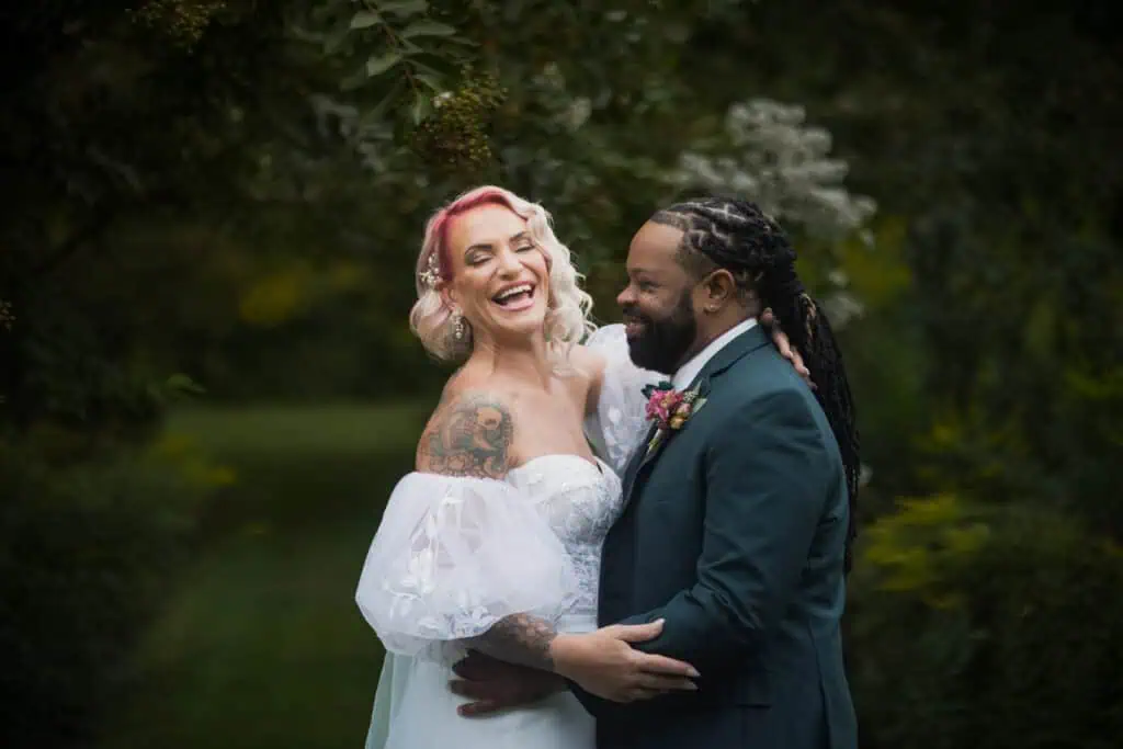 A joyful bride with pink hair and tattoos laughs with her groom, who has dreadlocks, in a lush garden setting.