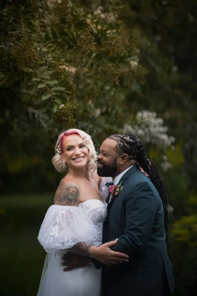 A joyful bride and groom embracing outdoors, surrounded by lush greenery, with the bride's tattoo visible on her arm.