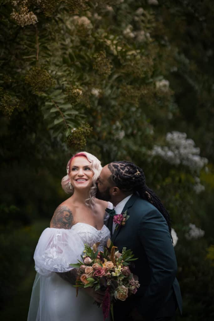 A bride with pink hair and a groom in a dark suit share a tender moment outdoors, surrounded by lush greenery and flowers.