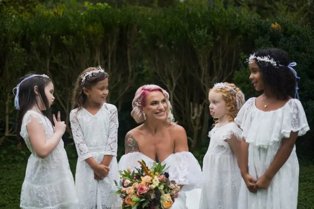 A bride with pink hair and tattoos smiles surrounded by four young bridesmaids in white floral dresses, all wearing floral crowns in a garden setting.