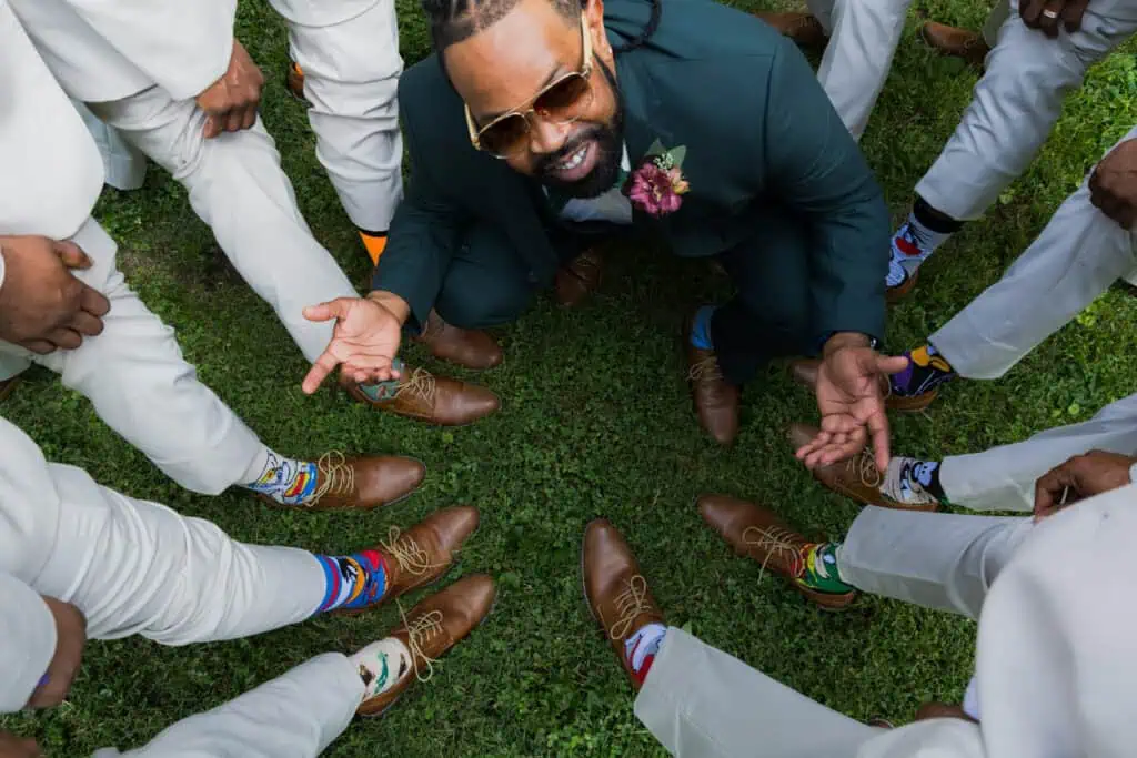 Groom and groomsmen showing off colorful socks and brown shoes in a circle on grass at a wedding.