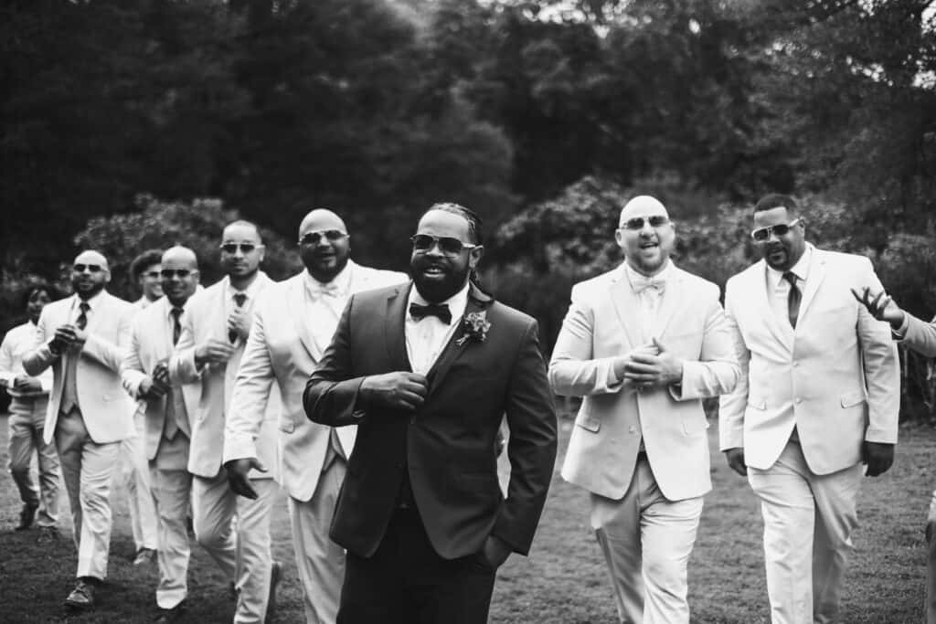 A black-and-white photo of a joyful groom in a black suit leading a group of groomsmen in white suits at a wedding, all smiling and walking outdoors.
