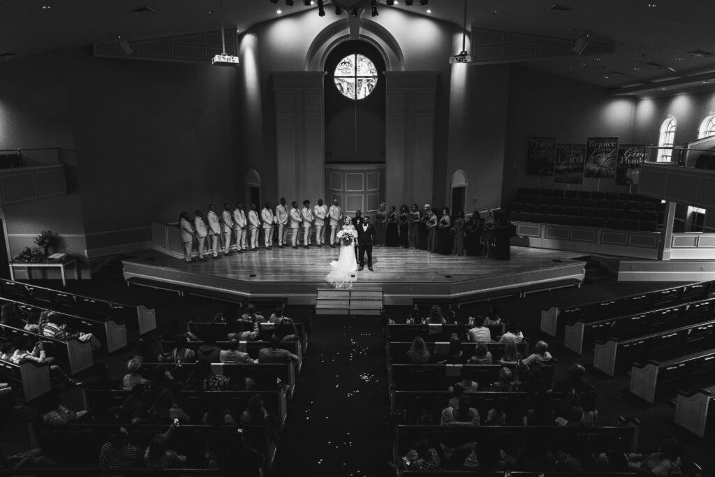 A black and white photo of a wedding ceremony in a church, with the couple standing at the altar and guests seated, viewed from the back of the church.
