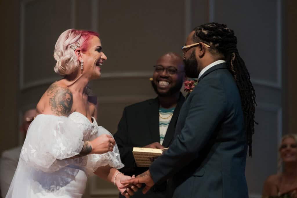 Bride with pink hair and tattoos and groom holding hands at the altar, with an officiant and guest smiling in the background.
