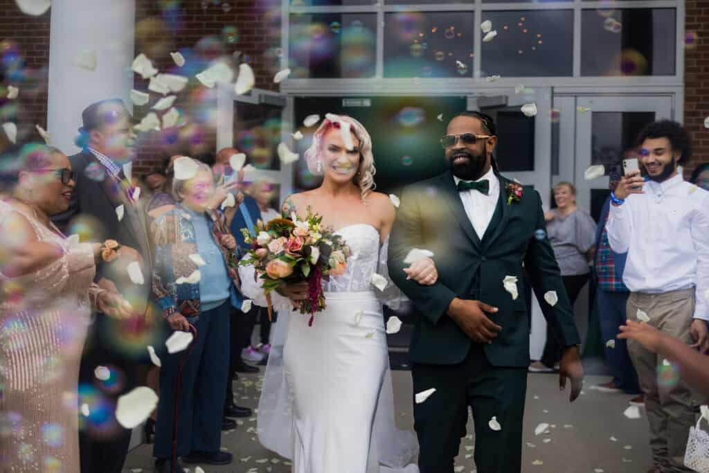 A newlywed couple walks joyfully through a crowd of guests throwing confetti outside a building.