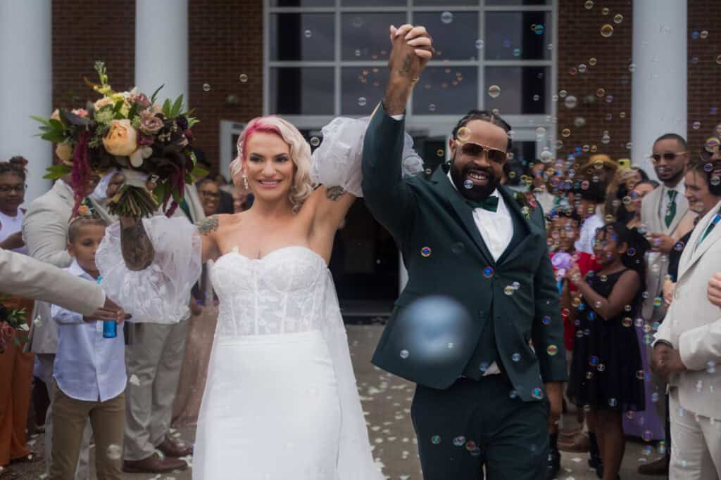 A joyful bride and groom exit a building while holding hands, surrounded by guests and bubbles, celebrating their wedding day.