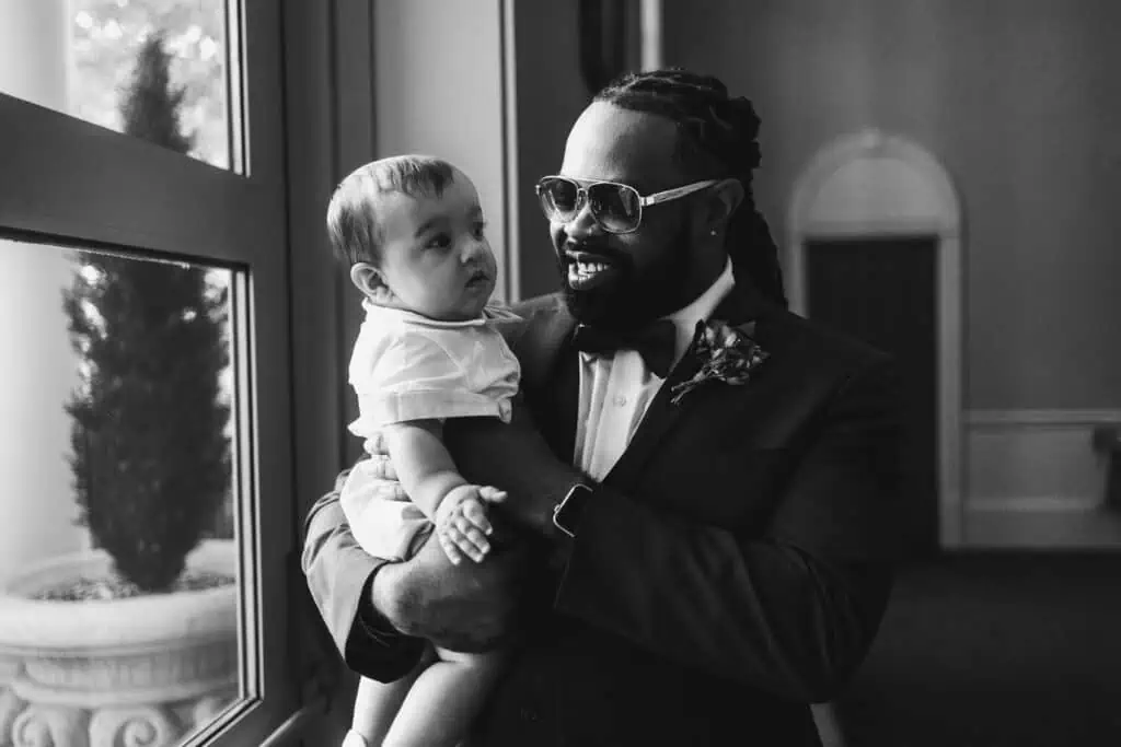 A man in a suit and sunglasses smiles while holding a baby near a window in a monochrome photograph.