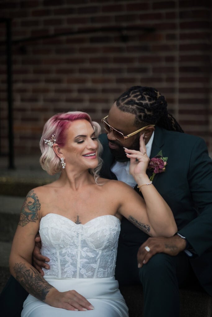 A bride with pink hair and a tattooed arm smiles at her groom, who is adjusting her earring, both seated against a brick wall.