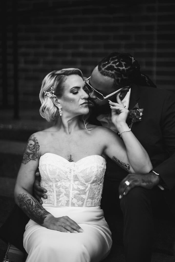 A groom in sunglasses kisses the forehead of a bride with tattoos, both dressed in wedding attire, sitting on steps against a brick wall.