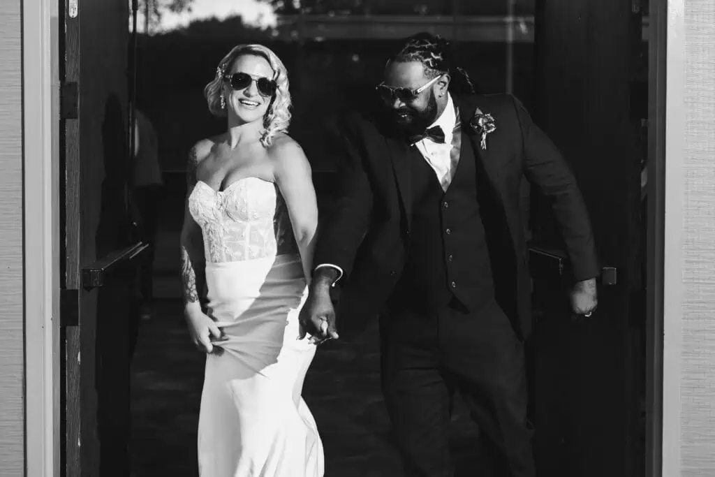A joyful bride and groom, hand-in-hand, exiting a building with the bride wearing a white strapless dress and the groom in a dark suit and sunglasses.