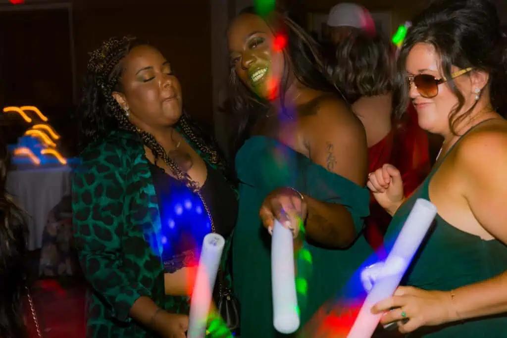 Three women enjoying a lively dance party with colorful lights and glowing wands, smiling and dancing together.