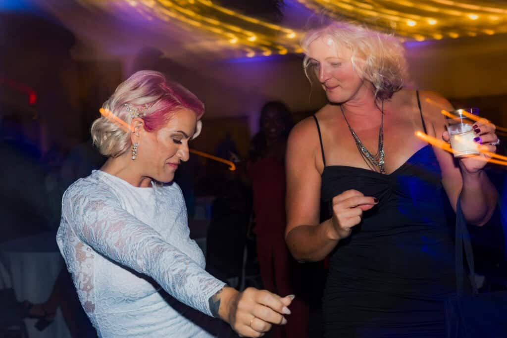 Two women dancing joyfully at a party with colorful lighting and one holding a drink.